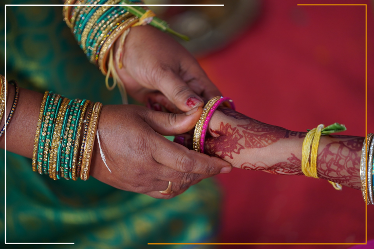 This Image appears to depict a close-up of two hands engaged in a traditional activity. One hand features intricate henna designs and is being adorned with a bangle by the other hand, which also wears multiple bangles. The vibrant red background adds to the cultural ambiance, suggesting a significant event—possibly related to marriage or festival celebrations. 🌺🤝💍