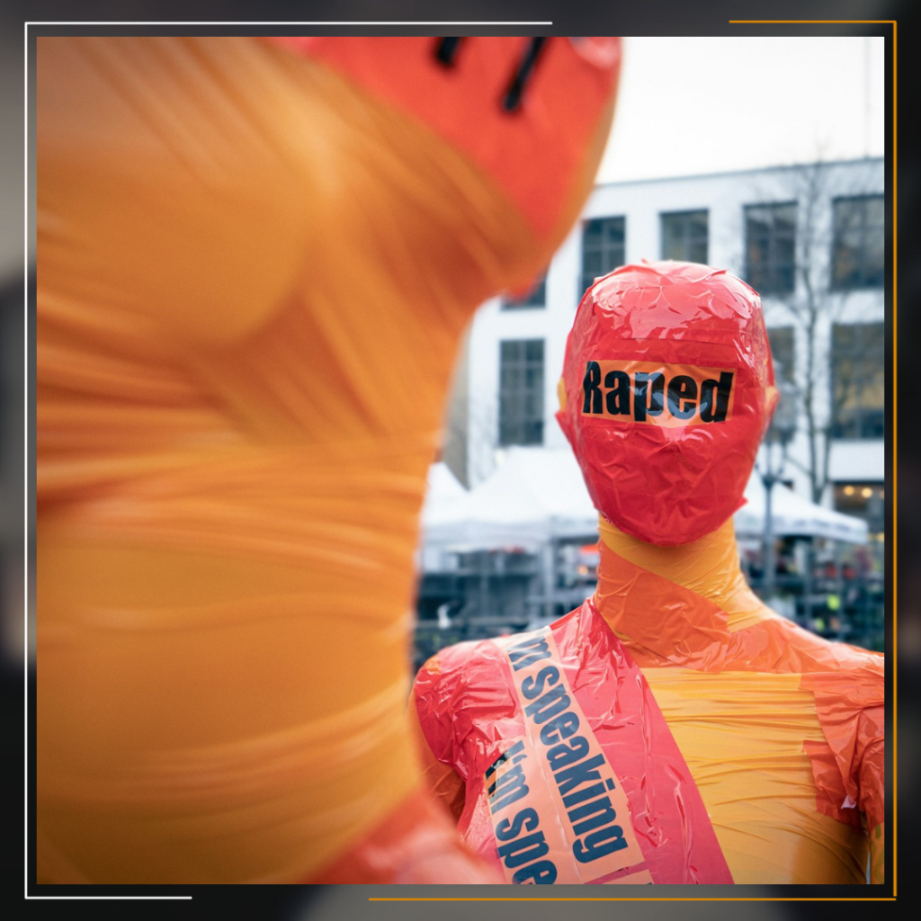 Miracles Law Firm It appears to depict two humanoid figures made from inflated plastic. The one in the foreground has a red head labeled “Rated,” while a larger figure stands in the blurred background. The red figure bears the text “is speaking till sep” on its torso. The overall scene seems to be an urban street.